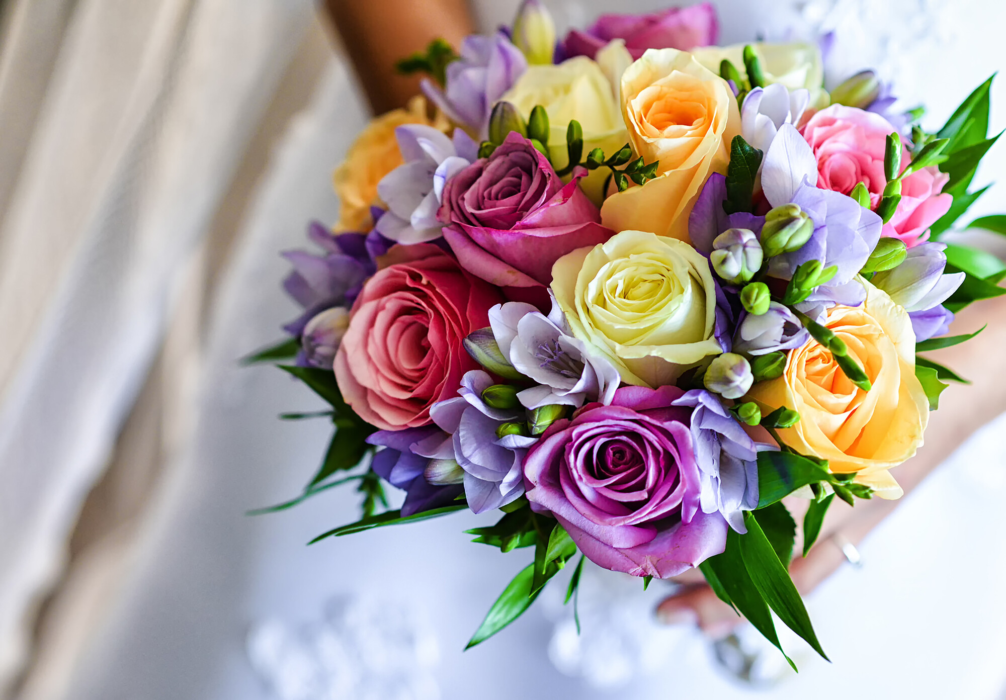 Bouquet da sposa con rose rosa, gialle e lilla, fresie viola e foglie verdi, tenuto da una donna in abito bianco