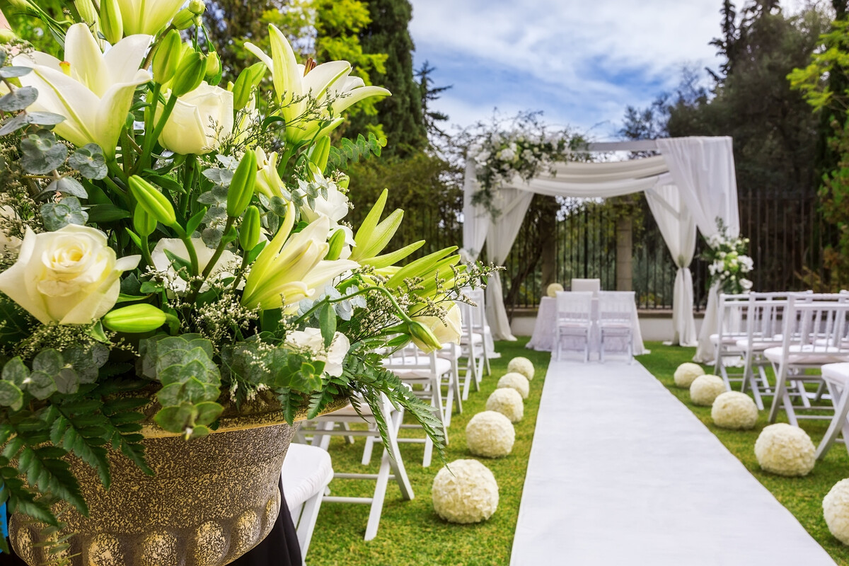 Gigli e rose bianche in vaso dorato, tappeto chiaro tra sedie candide e sfere floreali verso un gazebo drappeggiato